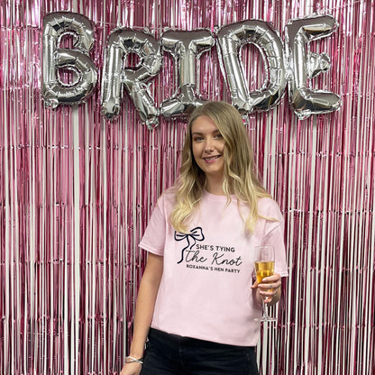 Woman holding a glass of champagne in front of 'BRIDE' balloons and pink tinsel curtain.