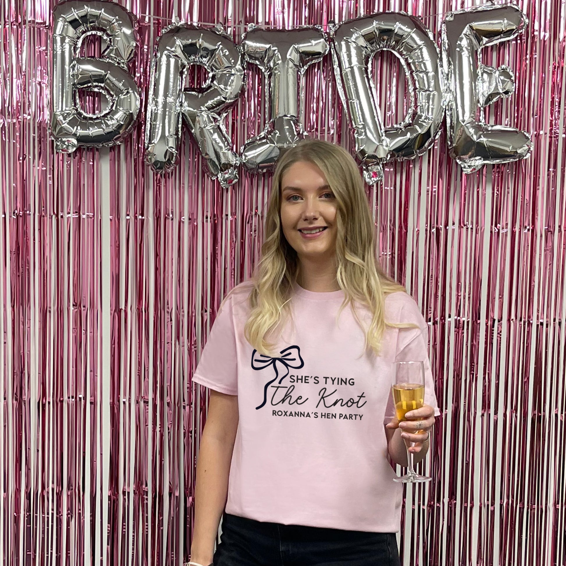 Woman in a pink t-shirt with 'She's Tying the Knot' text, holding a glass, in front of 'Bride' balloons and pink streamers.
