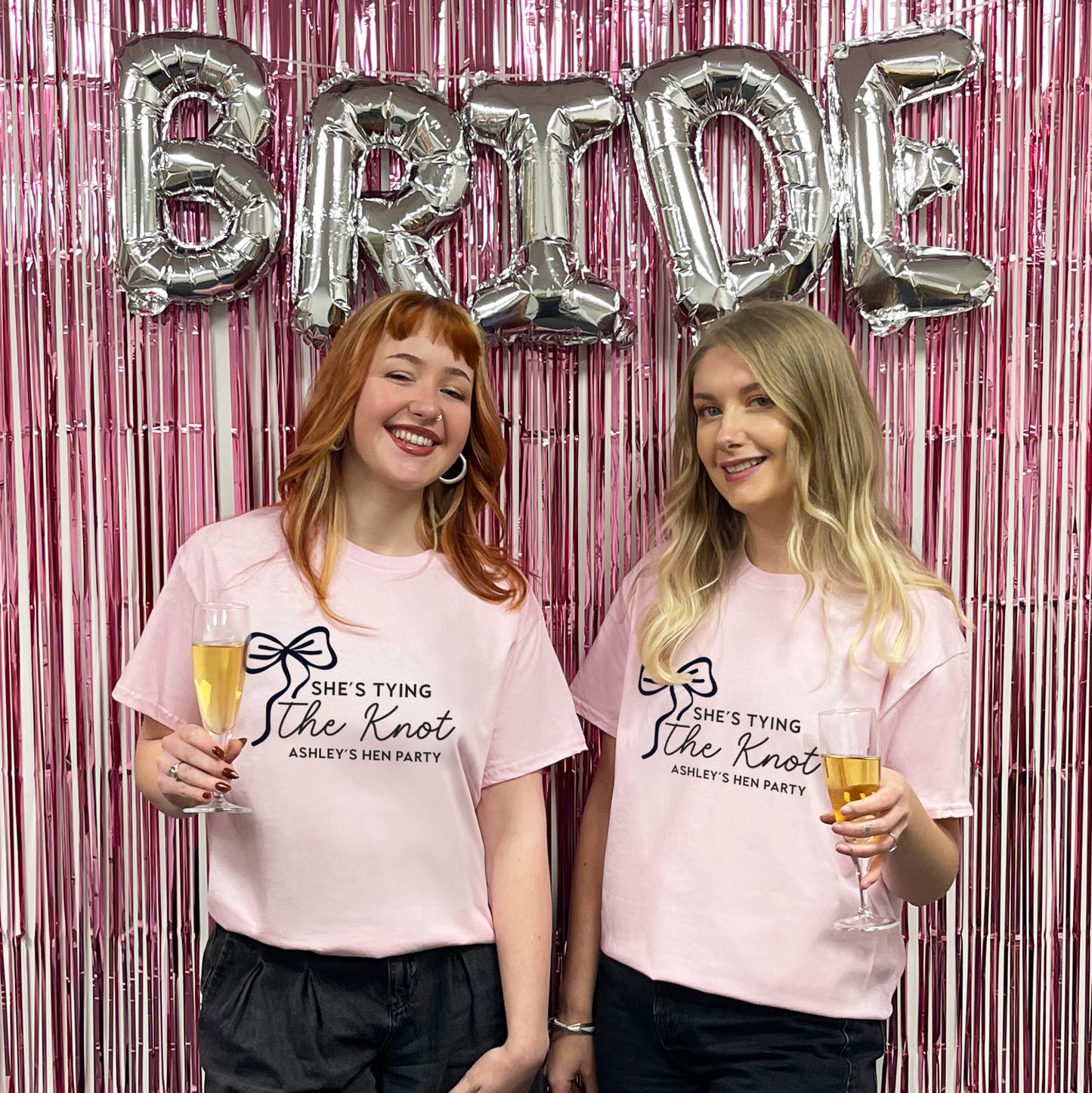 Two women in pink t-shirts with a hen party message, standing in front of 'BRIDE' balloons and a pink curtain.