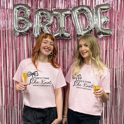 Two women in pink t-shirts with a hen party message, standing in front of 'BRIDE' balloons and a pink curtain.