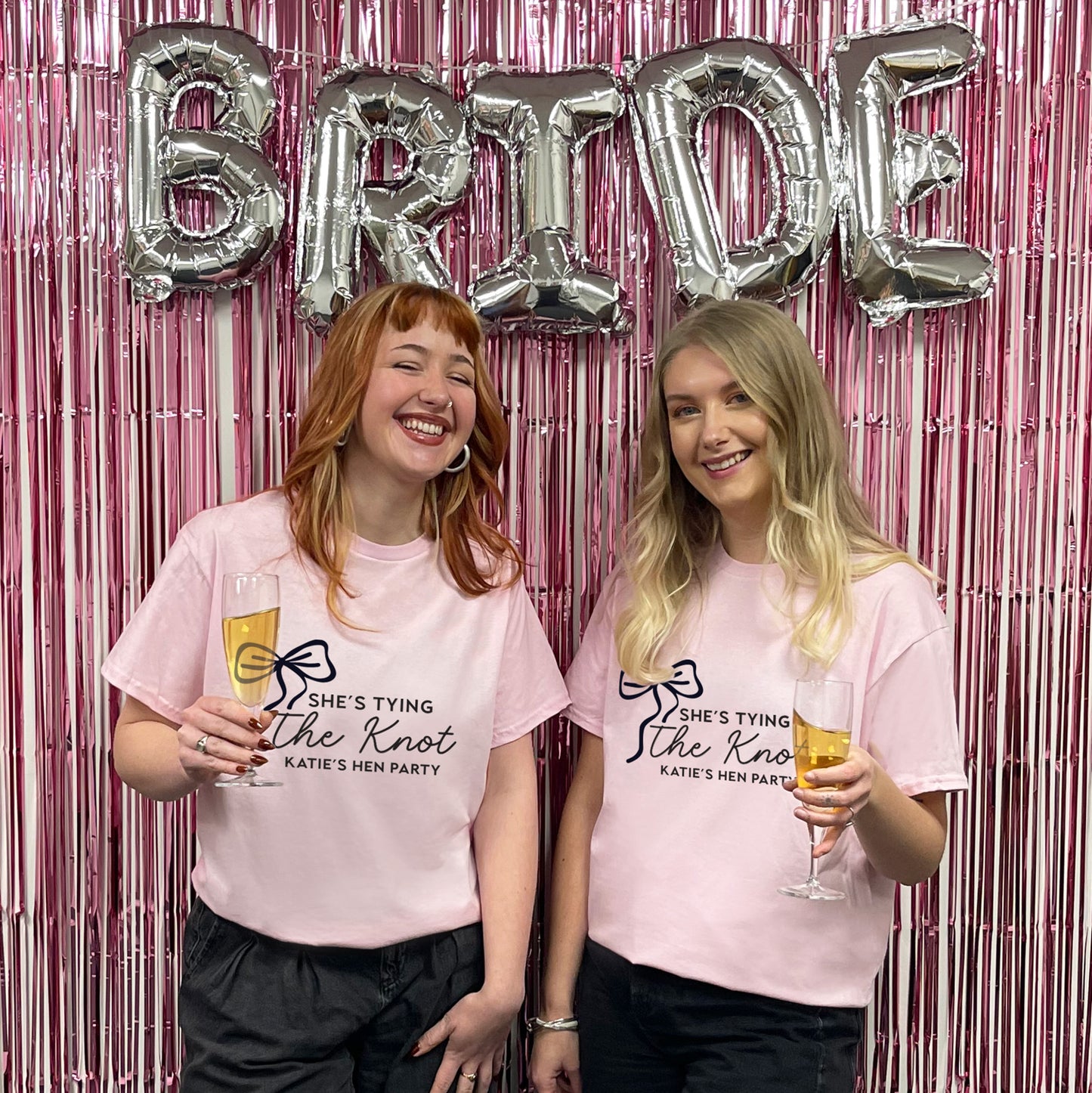Two women in pink t-shirts with hen party text, holding drinks in front of a 'BRIDE' balloon backdrop.