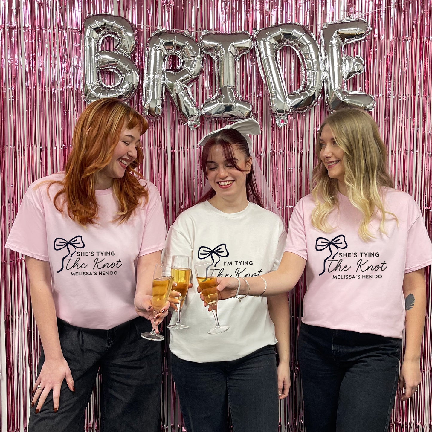 Three women wearing t-shirts with 'She's tying the knot' text, holding drinks in front of a pink 'BRIDE' balloon backdrop.