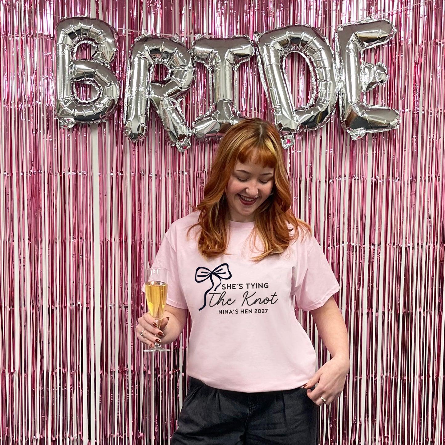 Woman holding a glass of champagne in front of 'BRIDE' balloons and pink streamers.