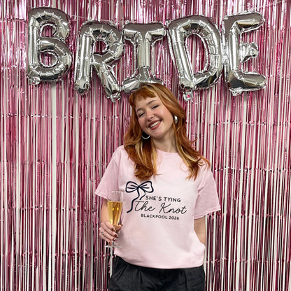 Woman holding a glass of champagne in front of 'BRIDE' balloons and a pink t-shirt with text.