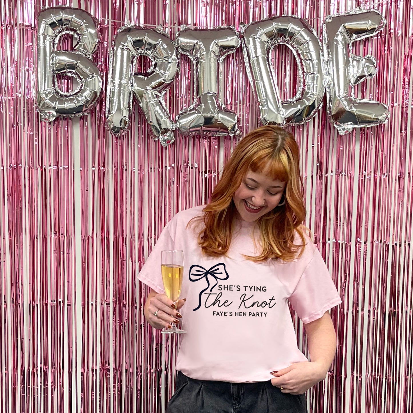 Woman holding a glass of champagne with 'BRIDE' balloons and pink tinsel in the background