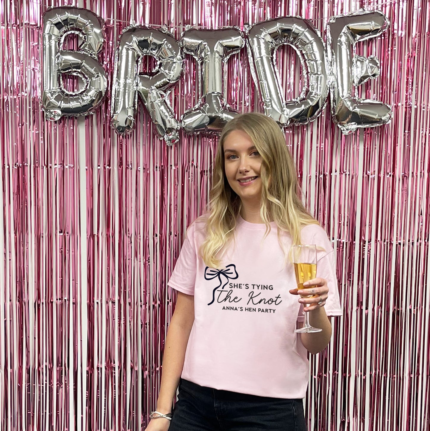 Woman in a pink t-shirt with 'She's Tying the Knot' text, holding a glass of champagne, in front of a 'BRIDE' balloon display.