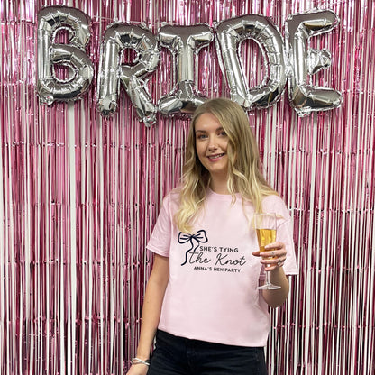 Woman in a pink t-shirt with 'She's Tying the Knot' text, holding a glass of champagne, in front of a 'BRIDE' balloon display.