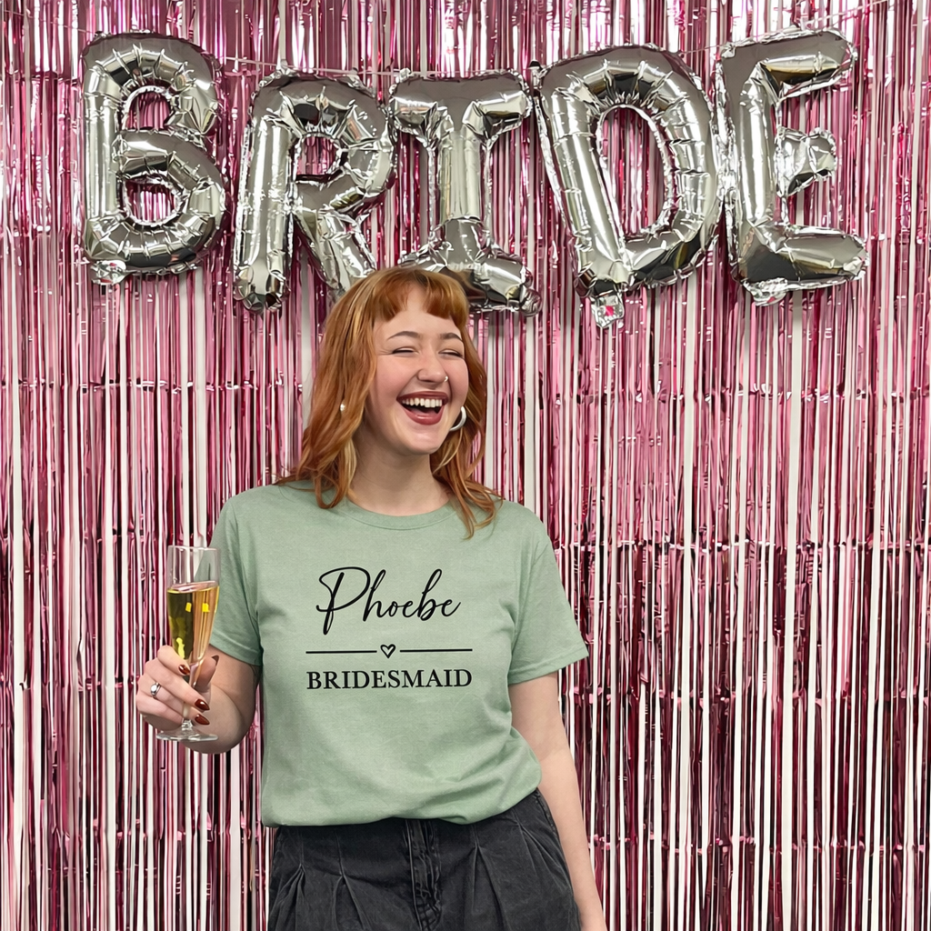 Woman in a 'Phoebe Bridesmaid' shirt holding a glass, with 'BRIDE' balloons in the background.